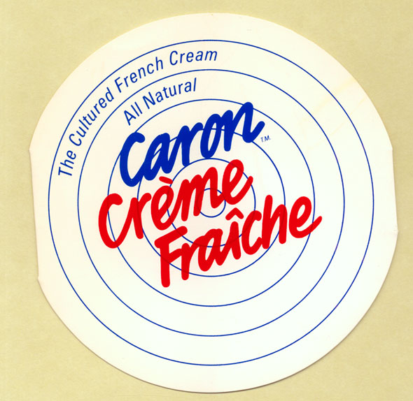 A photo of Circular white card with thin blue lines on it resembling a target. Text reads in blue and red: The Cultured French Cream, All Natural, Caron Crème Fraîche.  Caron Crème Fraîche is in a bold cursive font.