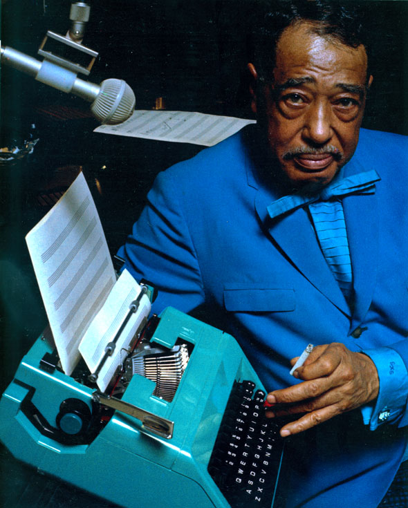 An old man in a bright blue suit ,near a microphone with music sheets in the background. holding a teal typewriter and hold a cigarette in his other hand. The typewriter has a music sheet set in place. 
