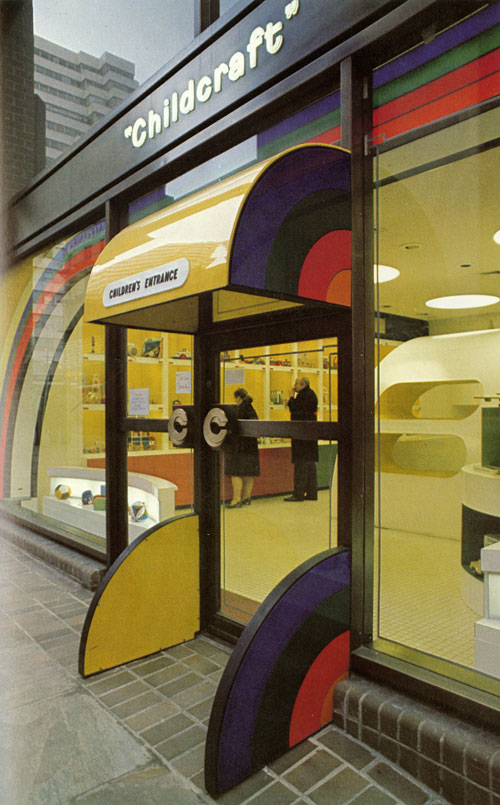 A close-up photo of the entrance of the location. The is two sets of doors, one for entering and one for exiting. The entrance has a bright yellow rigid storefront awning. On the side of the awning is a quarter circle rainbow pattern. There is a pair of small quarter-circle rainbow walls on the sides of the entrance door. You can see in the inside, two adults are looking at the cubbies that is filled with toys and other kid-like things.