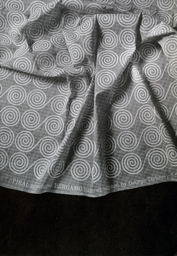 A photo of a grey thin cloth that has patterns of white circular spirals. At the bottom of the cloth "SPIRAL an exclusive BERGAMO handprint design by George Tscherny" is printed in white text. The cloth is scrunched in the middle and is not covering the whole image. Below the cloth is a black space.
