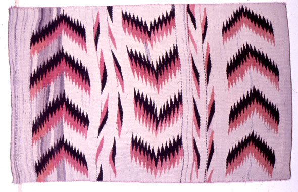 A hand-made rug. The rug is rectangular with a light lavender base color. There are designs that look like the tip of an arrow colored dark purple and salmon pink in a row at the left, middle, and right side of the rug. Between the arrow designs is another design that is smaller and made with the same color. 