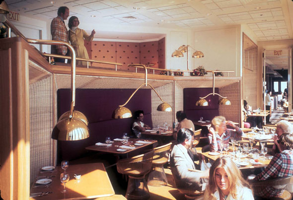 A photo of inside a restaurant. The restaurant has two levels. On the top level there is a couple standing looking down at the bottom level. The bottom level is the dinning room, that is filled with tables and chairs. 3 tables are occupied with costumers eating. The restaurant has a sunlight coming from the windows, and it has gold small lamps hanging form the bars from the top level. 