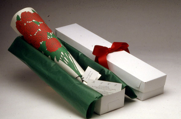 A white pencil box containing a rolled sheet of a paper, displaying a rose bouquet print. The box interior is layered with green wrapping paper.