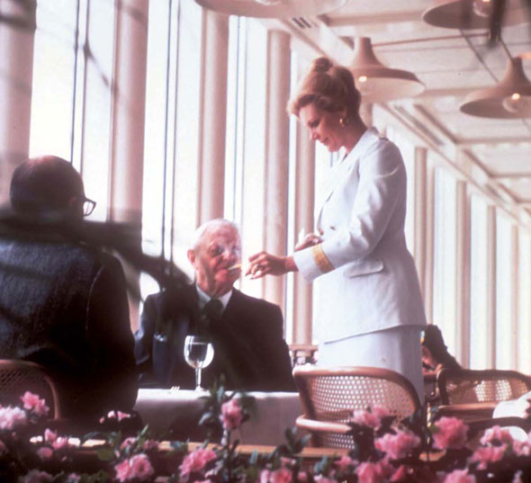 A photo of two old white men sitting near a window in a pristine white fancy restaurant. The oldest of the two men has a cigarette in his mouth. A woman in a clean cute white blazer and skirt outfit is standing and lighting the old man's cigarette. 
