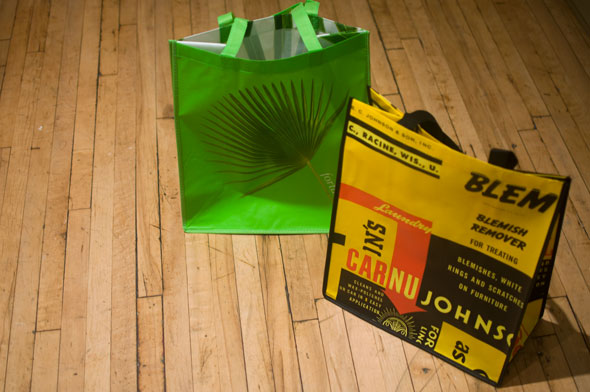 A photo of Two reuseable shopping bags on the floor; one is green with a dark green palm leaf on it, the other yellow and black with dense text and a red arrow.