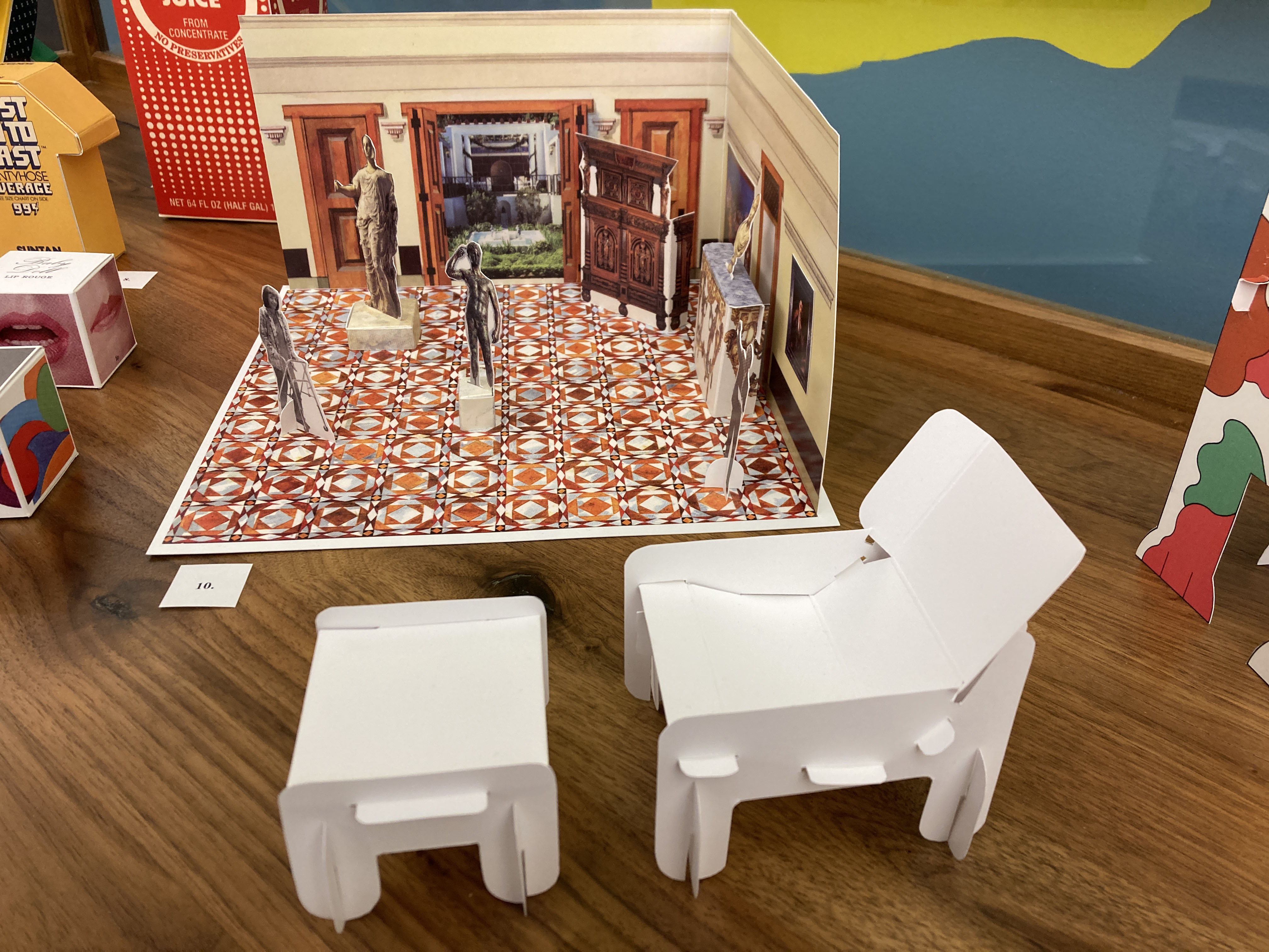 In the background, a model of a musem room with paper cut-out art and visitors. In the foreground, a scale model of a chair and footstool made of punched-out paper