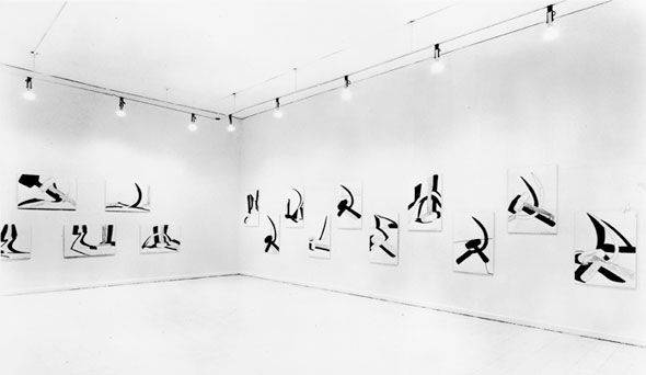 A photo of a plain room withe white floors, ceiling, and flooring. Mounted on the wall are black and white artwork hung in a zig-zag pattern. 