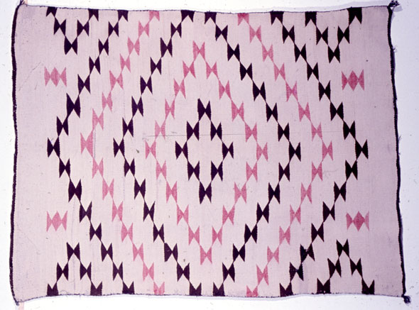 A hand-made rug. The left and right side have a thin purple border. Each corner of the rug has a small purple tassel. The rug has pastel pink base color and has a complex design of triangles made with a darker pink and purple.