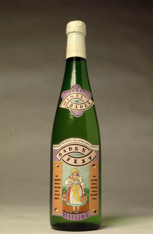 A photo of a sealed green champagne bottle. On the upper half of the bottle is a complex design sticker with "The Wonderful Wines of Baden" on it. The label of the bottle has a colorful illustration of a country maiden with her hand on her hip and carrying a basket. Above the illustration "Baden Fest" in printed on top on a peach banner. Underneath the illustration  "REISLING" in printed on a small purple banner. 