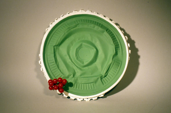 A photo of a sculpture. It's a outdated hub cap with its center crushed in. The border from the underside of the hubcap is pushed outward and is painted white with the front border of the hub cap. Rest of the hub cap is painted a lime green. On the bottom left small plastic grapes adorn the hub cap. The hub cap is transformed into a fancy green sun hat. 