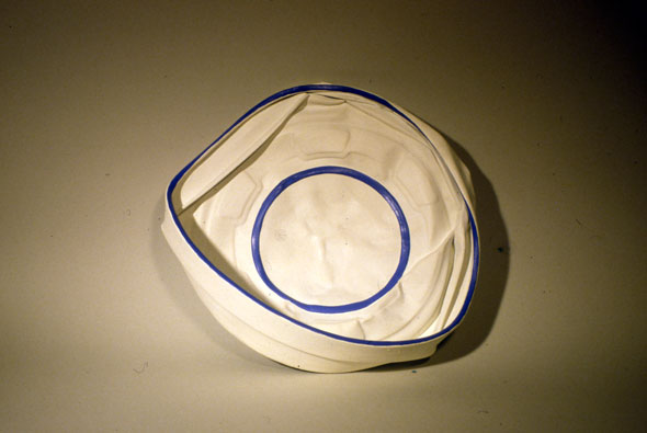 A photo of a sculpture. It's a outdated hub cap with its outer border crushed towards the center, giving it folds. The outline of the hub cap's border is painted with a thin blue line, so it the circle drawn in the center of the hub cap. The rest of the hub cap is painted white. The hub cap is transformed into a sailor hat. 