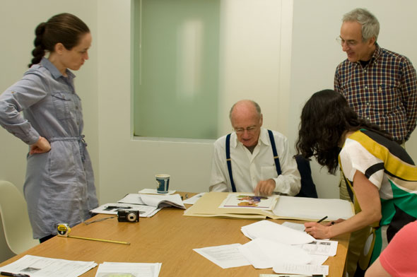 A photo of 4 people gathered around a table that has paper scattered. James McMullan is seated, in the middle of everyone who is standing. He is looking over his past work. 