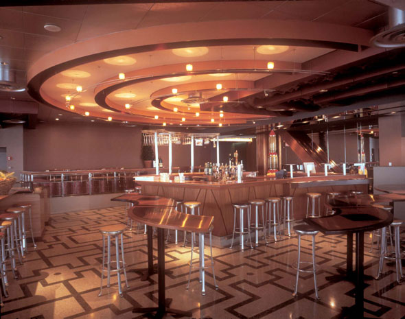 A photo of inside a restaurant. It's the bar section of the restaurant with only high cherry wooden tables and high grey stools. At the center is the bar. The bar is small and has a counter so people can drink there. The bar has a very small display of alcohol in the center. The ceiling right above the bar are arches that spread outward to half the room. The light on it have a very warm color. 