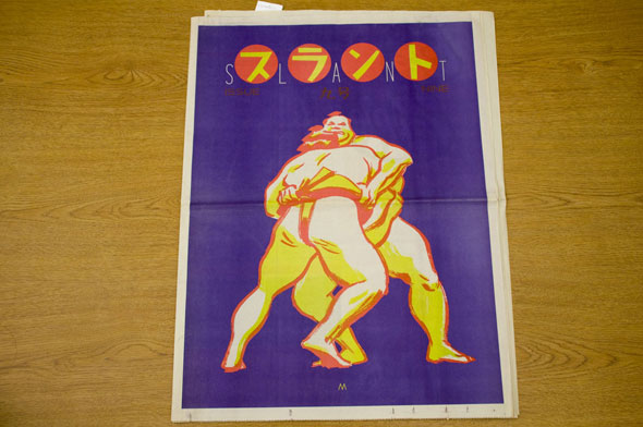 A newspaper for "Slant". The front page is two sumo wrestlers wrestling in a bright blue background. The Sumos are colored white and yellow with a orange outline. Above the sumo is yellow Japanese calligraphy in small orange circles. Next to each circle are thin white letters that spell out "Slant".