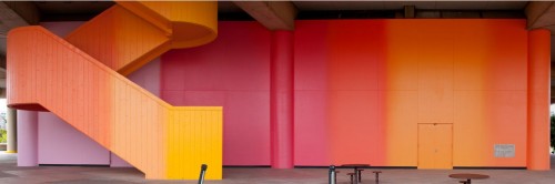 A photo of a wall and staircase. The wall and staircase has a gradual vertical transition across it with different variation of colors such as orange, pink, red, and light pink.