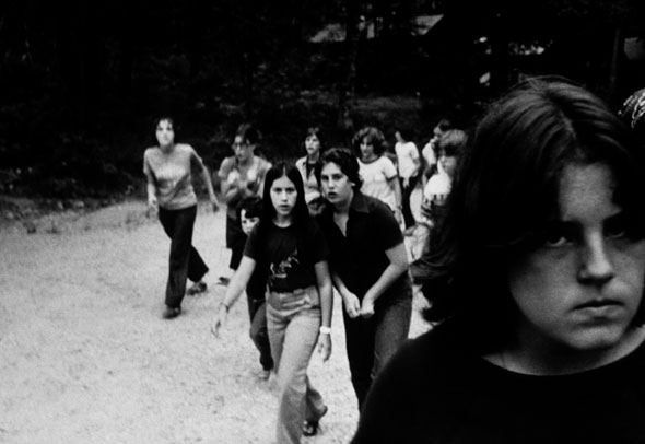 Black and white photograph of a children walking outside; a person in the foreground is close-up and looking into the camera.