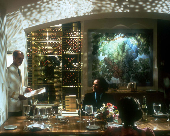 A photo of inside a restaurant. The dining room has a big wine stack on the walls and has a big painting of wine grapes next to it in the background. In the foreground there is along table with only two people sitting. They are being waited by a waiter in a clean cut white blazer uniform. The room is quite dark and has shadows that resembles shadows from a tree, with a few rays of lights seeping through the holes of leaves. 