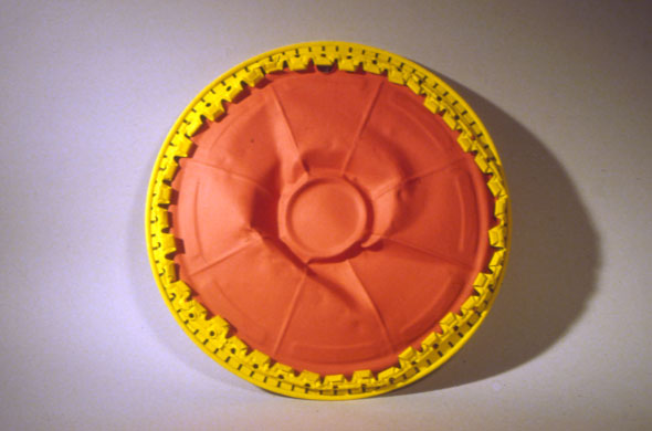 A photo of a sculpture. It's a outdated hub cap with its center pushed outward. The hub cap is painted red and adorned with a yellow metal going around the hub cap. 