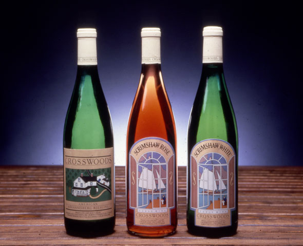 A photo of 3 wine bottles. The leftmost green bottle is 'Cross Woods' Wines. The label is an illustration of a white suburban house on a green field. The middle clear bottle  is " Scrismshaw Rose". The label is an illustration of a wooden boat with white sails viewed from insides a window. The rightmost green bottle is "Scrimshaw White". The label is an illustration of a wooden boat with white sails viewed from insides a window.