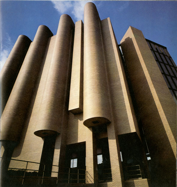 A photo of a worm-eye view of a building. The building is made of brown bricks. The entrance has 4 thin cylinder wall design popping out of the plain flat wall.