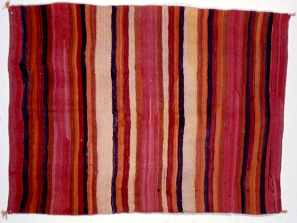 A hand-made rug. The rug is medium size with the top and bottom slightly longer than the sides. At each corner there is a small tassel. The rug has vertical stripes made of different hues of red, orange, and purple.