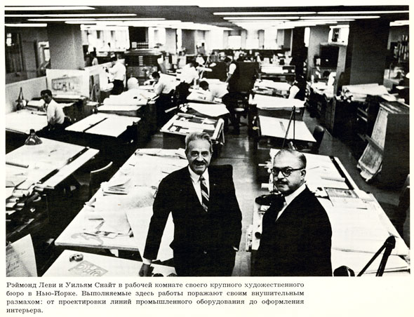 Black and white photo of an office of graphic designers at their elevated tables. Two men in the foreground are standing up and looking at the camera