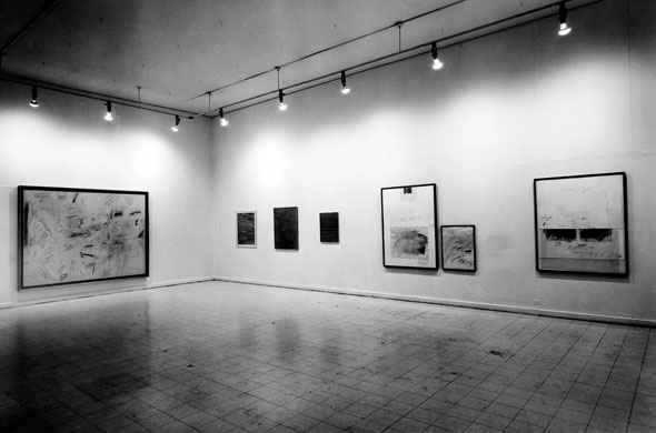 Black and white photo of the interior of an art gallery, showing 7 paintings