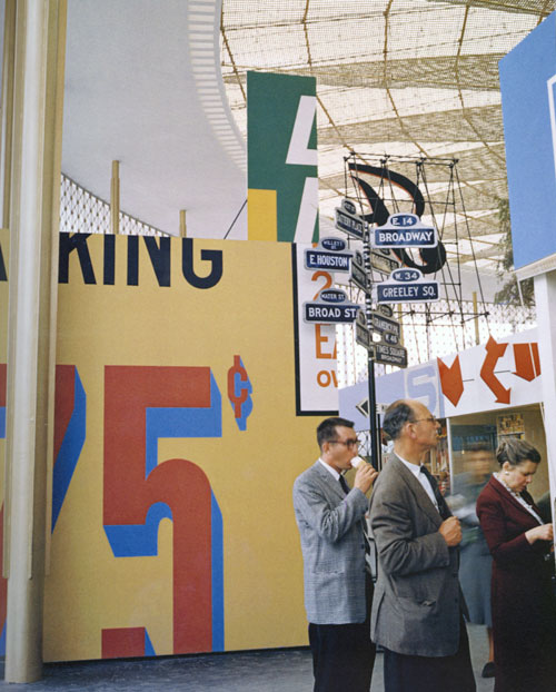 Color photograph of a street scene, showing the large typography on signs; Three suited people, two men and a woman, are visible in the foreground.