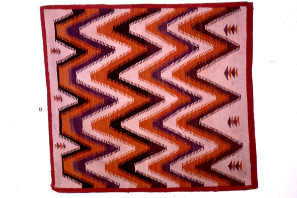 A hand-made rug laid out flat. The rug is small and square. It has a red boarder surround a light pink design that is filled with black, orange, and purple zig-zag lines going across vertically. 