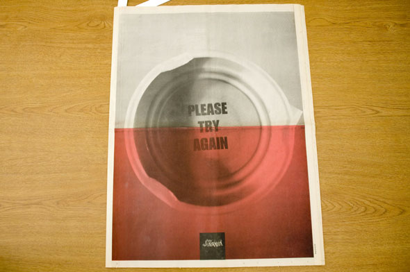 A newspaper for "Slant". The front/back cover is a photo of the underside of a bottle cap that has "Please Try Again" in bolded black text. The half of the image has a light red hue overlaid on it. 