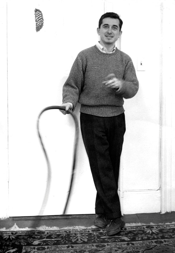 Black and white full body photo of a man in a sweater standing against a white wall