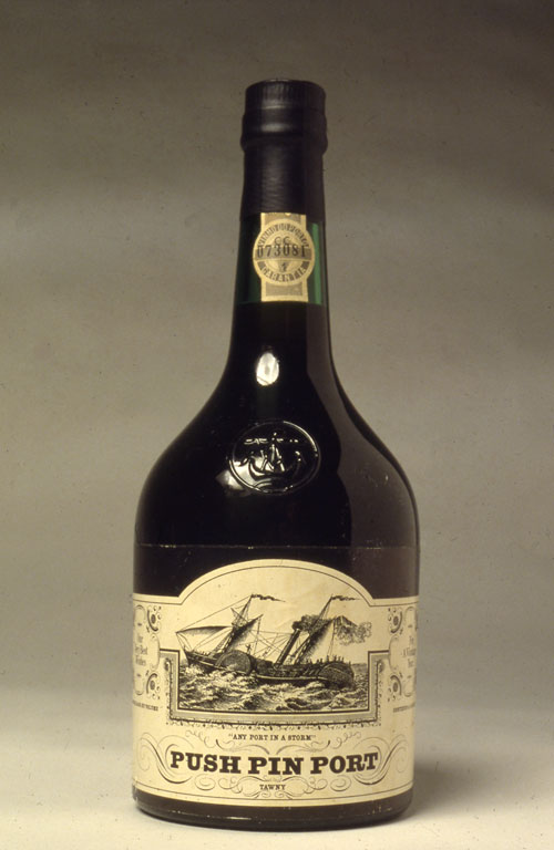 A photo of a short and stout alcohol bottle.The label for the bottle in a black and white illustration of a big wooden boat sailing on the sea. Below is "Any Port n a Storm" in very small think text and underneath is "Push Pin Port" In thick bold black text. The rest of the label has small fancy details. 