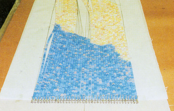 A photo of the process of putting together the beaded curtain. A line of strings that all start off being filled with blue beads then goes to light yellow beads and a part of an outline of a clouds begins to form down on the strings.