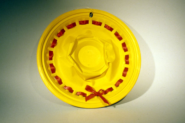 A photo of a sculpture. It's a outdated hub cap with its center crushed in. It's painted canary yellow and adorned with a red ribbon going around, through the small holes of the hub cap. The hub cap is transformed into a yellow sun hat.