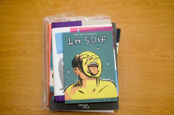 Photo of a Stack of books on a table; top reads La Soif and shows a yellow man sticking his tongue out to catch white droplets against a teal background