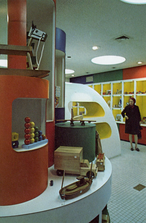 A photo of the back of the location. A geometrical colorful set of large and small cylinders with holes served as shelfs. The cylinder shelfs are on top of a white circular table that is covered in toys. The floor is tiny white swore tiles. The lighting on the ceiling are big white circular lights. And the wall of the cubbies is minimalistic yet colorful. The wall is a rainbow stripped design and the white cubbies has a bright yellow inside the cubbies.