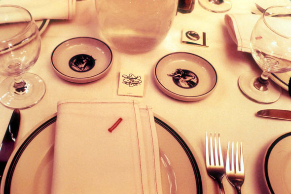 A photo of a fancy dining table. Each placement has a white clothed napkin folded normally on a small ordinary white plate. A knife is placed on the left and two forks on the right. In the center of the table is tiny plate, ashtrays, for each placement. The tiny ashtrays has a design of wood/farm animals.