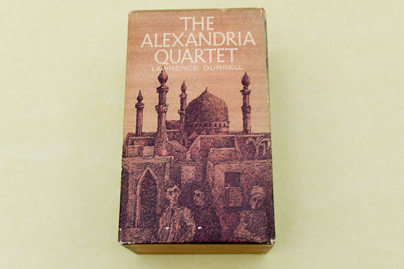 A photo of the book, "The Alexandria Quartet". The cover has the title printed on the top in thin white text. The background is a dusty brown sky. Below is a town of small houses and building in the middle adorned with detailed decorations. In the foreground, are 3 people who's form are lost with the dark sketches of the houses. 